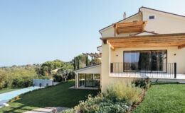 Modern villa with a spacious garden and swimming pool, featuring large windows and wooden pergola, surrounded by lush greenery and a clear blue sky. Ideal for luxury living and outdoor relaxation.