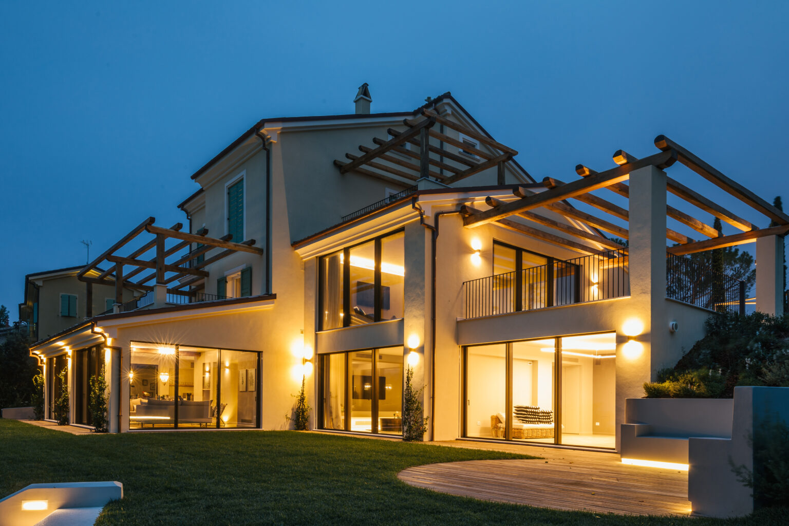 Modern luxury villa illuminated at dusk, featuring large glass windows, a spacious patio, and a stylish pergola. The well-manicured lawn adds to the elegant outdoor setting. | Only on SopranoVillas