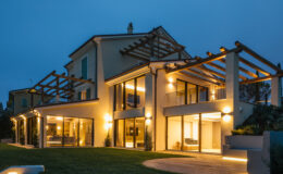 Modern luxury villa illuminated at dusk, featuring large glass windows, a spacious patio, and a stylish pergola. The well-manicured lawn adds to the elegant outdoor setting.