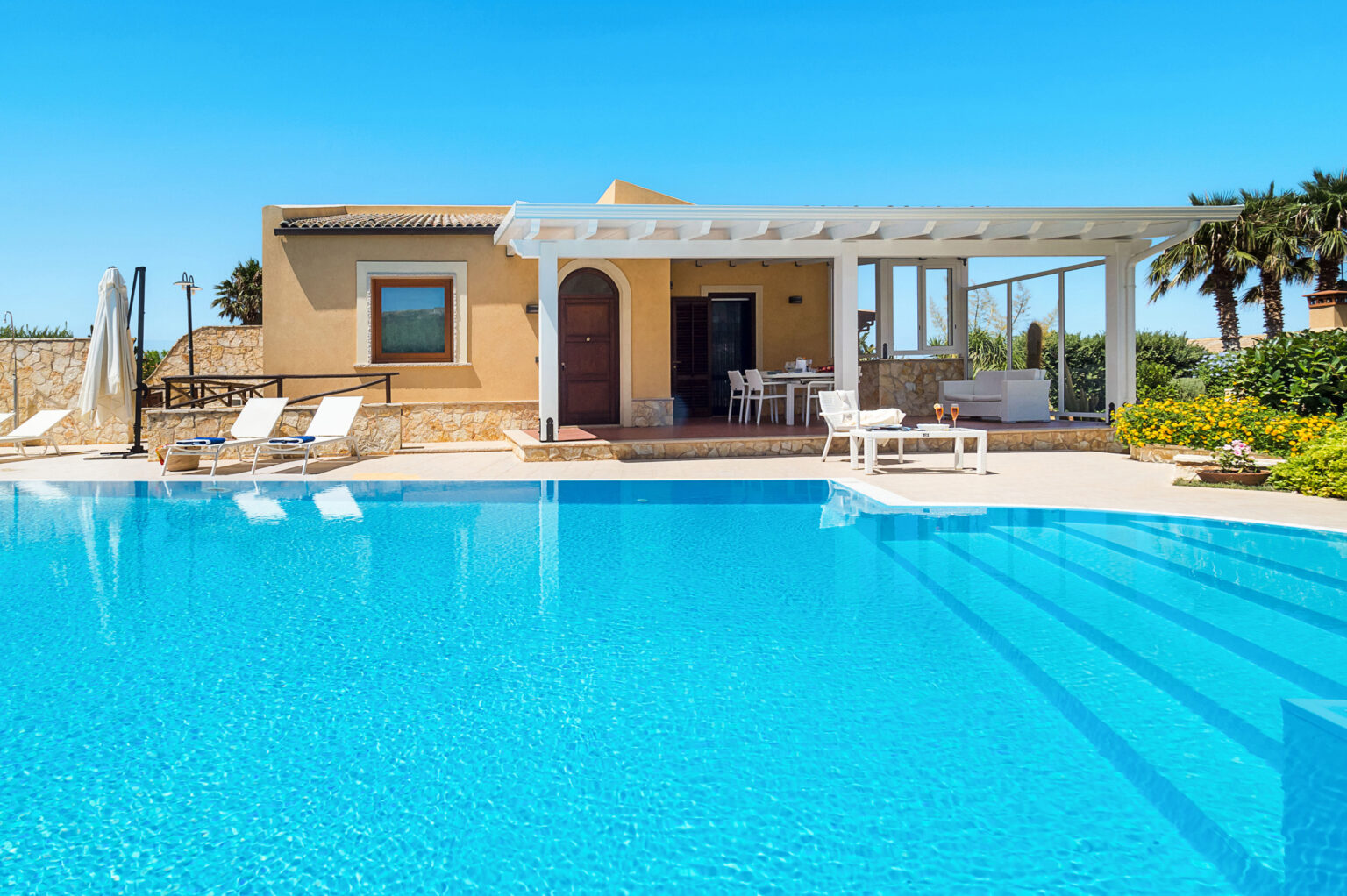 Modern villa with a swimming pool, surrounded by lush greenery and sunny skies. The outdoor space features lounge chairs, a shaded area, and a dining table, ideal for relaxation and entertaining. | Only on SopranoVillas