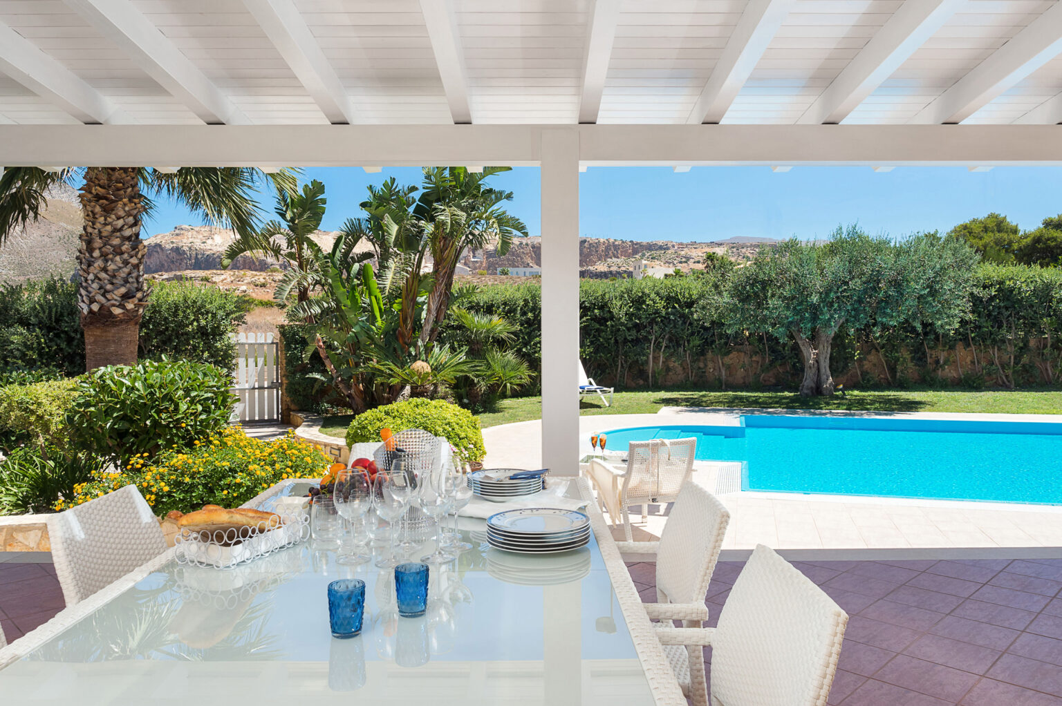 Outdoor dining area with a glass table set for a meal, featuring blue glasses and a view of a swimming pool and lush garden under a sunny sky. | Only on SopranoVillas
