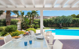 Outdoor dining area with a glass table set for a meal, featuring blue glasses and a view of a swimming pool and lush garden under a sunny sky.
