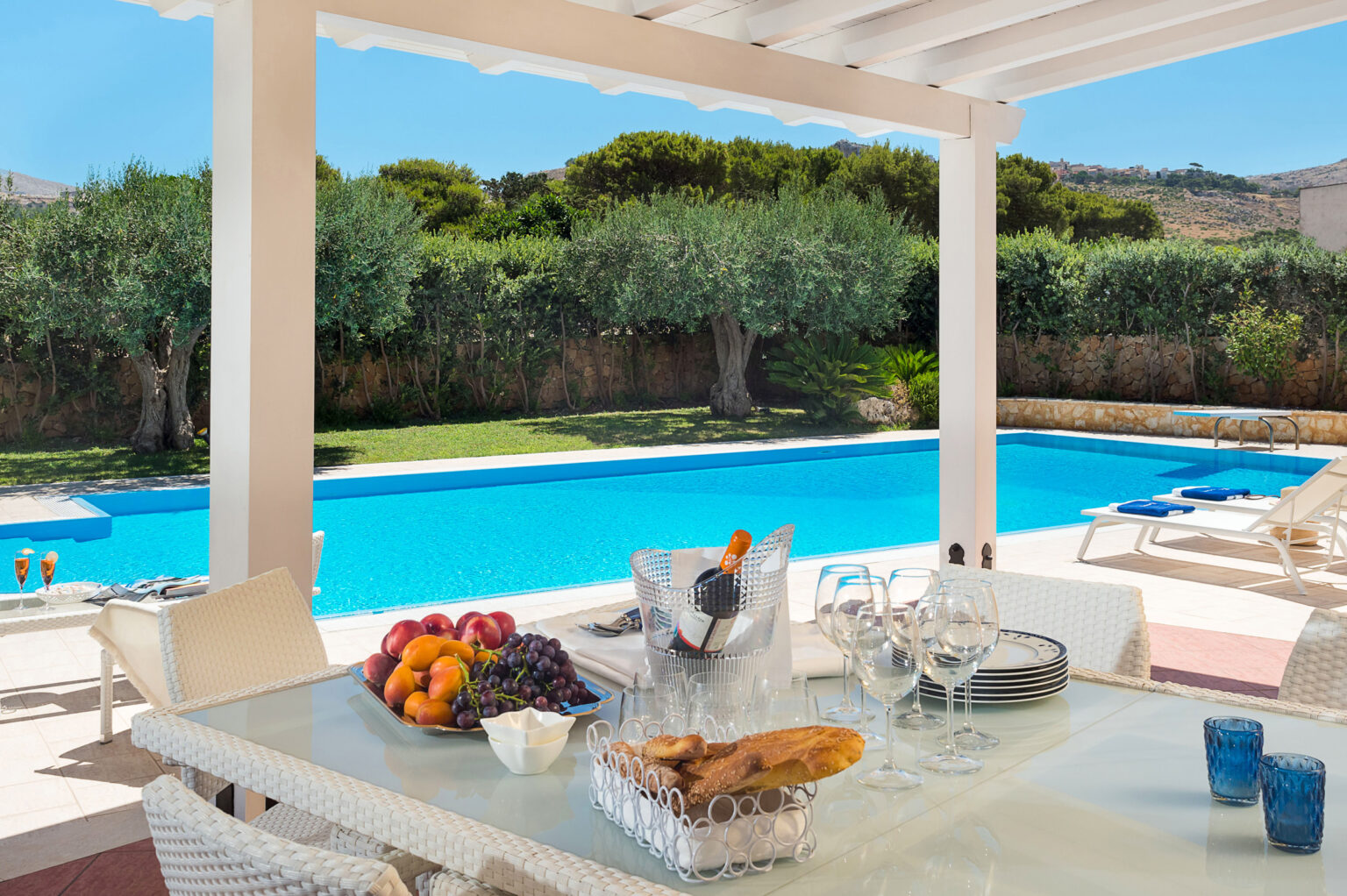 Elegant outdoor dining area featuring a table set with fresh fruits and a bottle of wine, overlooking a sparkling blue swimming pool surrounded by lush greenery. Ideal for relaxing summer gatherings and alfresco dining. | Only on SopranoVillas
