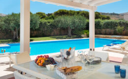 Elegant outdoor dining area featuring a table set with fresh fruits and a bottle of wine, overlooking a sparkling blue swimming pool surrounded by lush greenery. Ideal for relaxing summer gatherings and alfresco dining.
