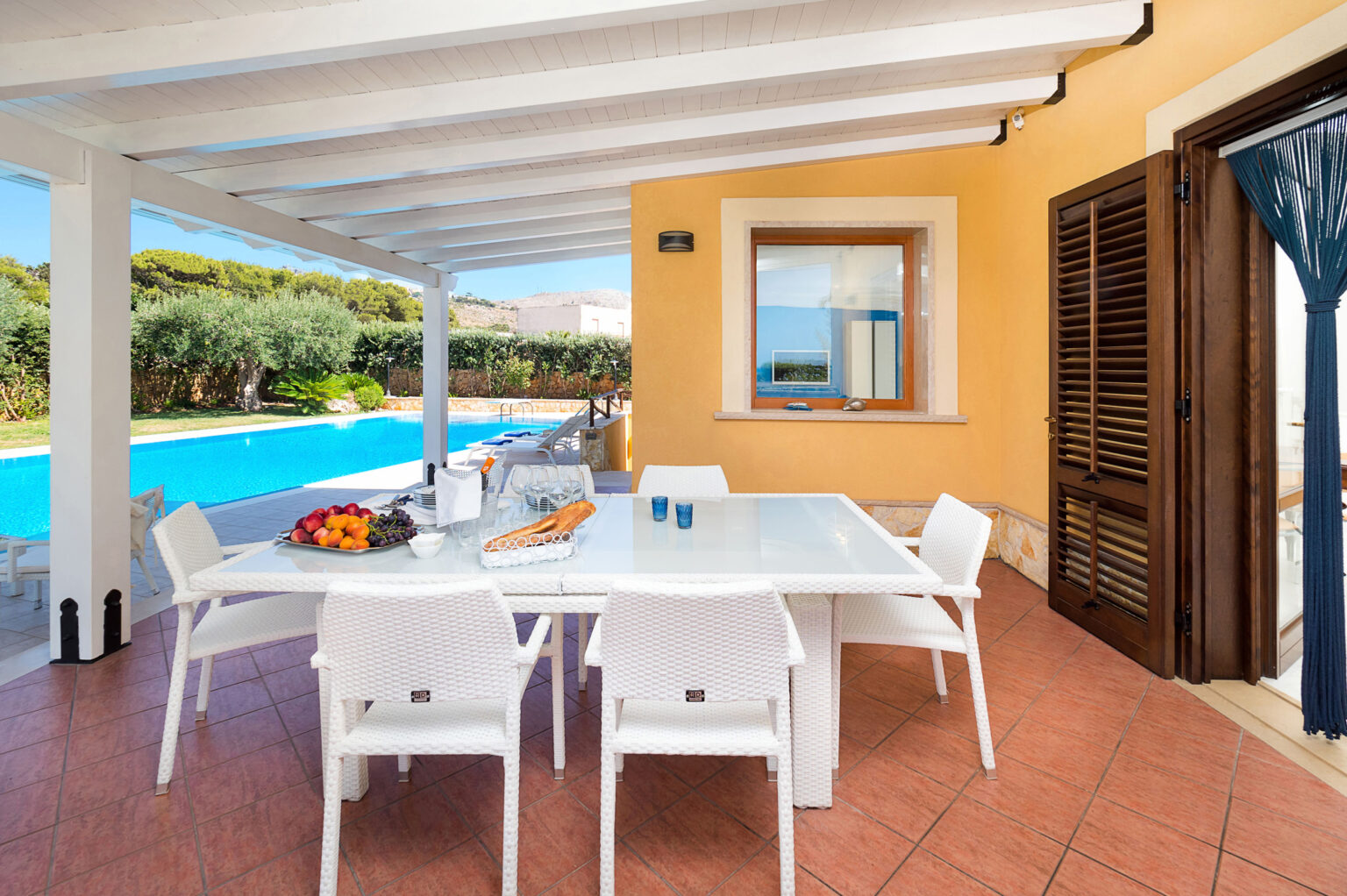 Outdoor dining area with a modern white table and chairs, overlooking a swimming pool surrounded by greenery, featuring a warm yellow wall and wooden shutters, ideal for relaxation and entertaining. | Only on SopranoVillas