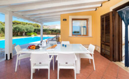 Outdoor dining area with a modern white table and chairs, overlooking a swimming pool surrounded by greenery, featuring a warm yellow wall and wooden shutters, ideal for relaxation and entertaining.