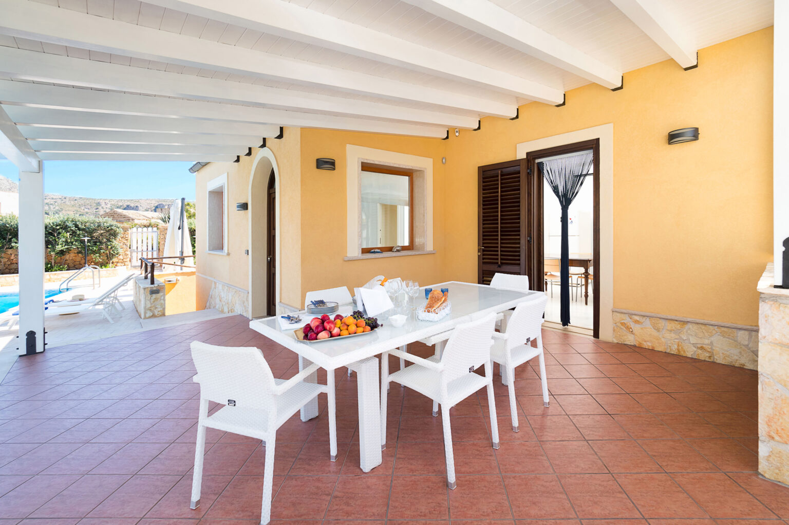 Bright outdoor dining area with a white table and chairs, featuring a fruit bowl, bread, and glasses. The space is surrounded by a sunny patio, with a view of a swimming pool and landscaped greenery. Ideal for relaxing meals in a Mediterranean setting. | Only on SopranoVillas