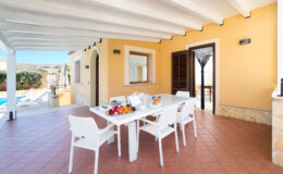 Bright outdoor dining area with a white table and chairs, featuring a fruit bowl, bread, and glasses. The space is surrounded by a sunny patio, with a view of a swimming pool and landscaped greenery. Ideal for relaxing meals in a Mediterranean setting.