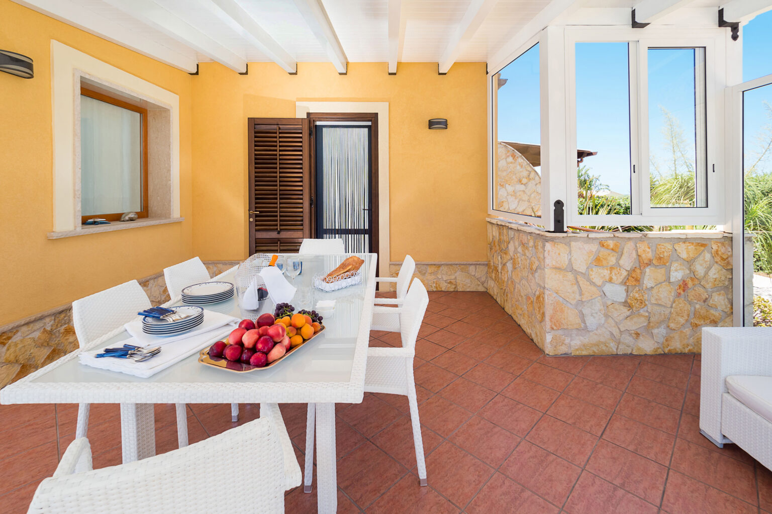 Bright and airy dining area with a large white table set for a meal, featuring fresh fruit and elegant tableware. The space is adorned with light-colored walls and large windows, offering a view of the outdoors. Perfect for al fresco dining in a sunny, inviting atmosphere. | Only on SopranoVillas
