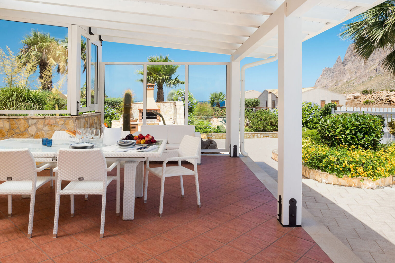 Bright outdoor dining area featuring a white wicker table and chairs, surrounded by lush greenery and palm trees, with a scenic view of mountains and the ocean in the background. Perfect for enjoying meals in a sunny, tranquil setting. | Only on SopranoVillas