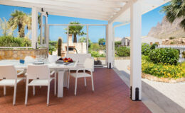 Bright outdoor dining area featuring a white wicker table and chairs, surrounded by lush greenery and palm trees, with a scenic view of mountains and the ocean in the background. Perfect for enjoying meals in a sunny, tranquil setting.