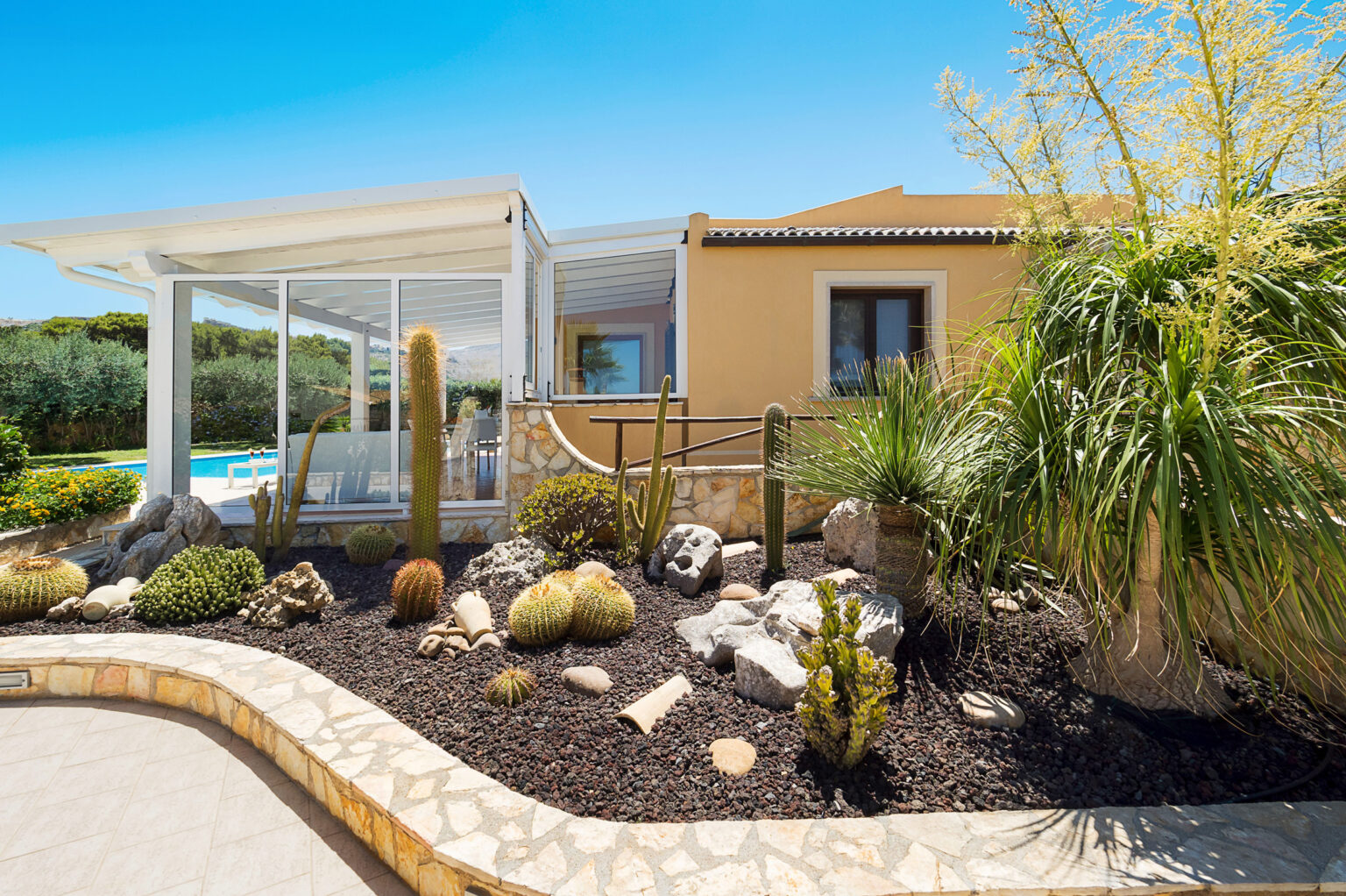 Modern villa exterior featuring a landscaped garden with cacti and tropical plants, a pool visible in the background, and a sunny blue sky. | Only on SopranoVillas