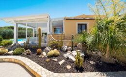 Modern villa exterior featuring a landscaped garden with cacti and tropical plants, a pool visible in the background, and a sunny blue sky.
