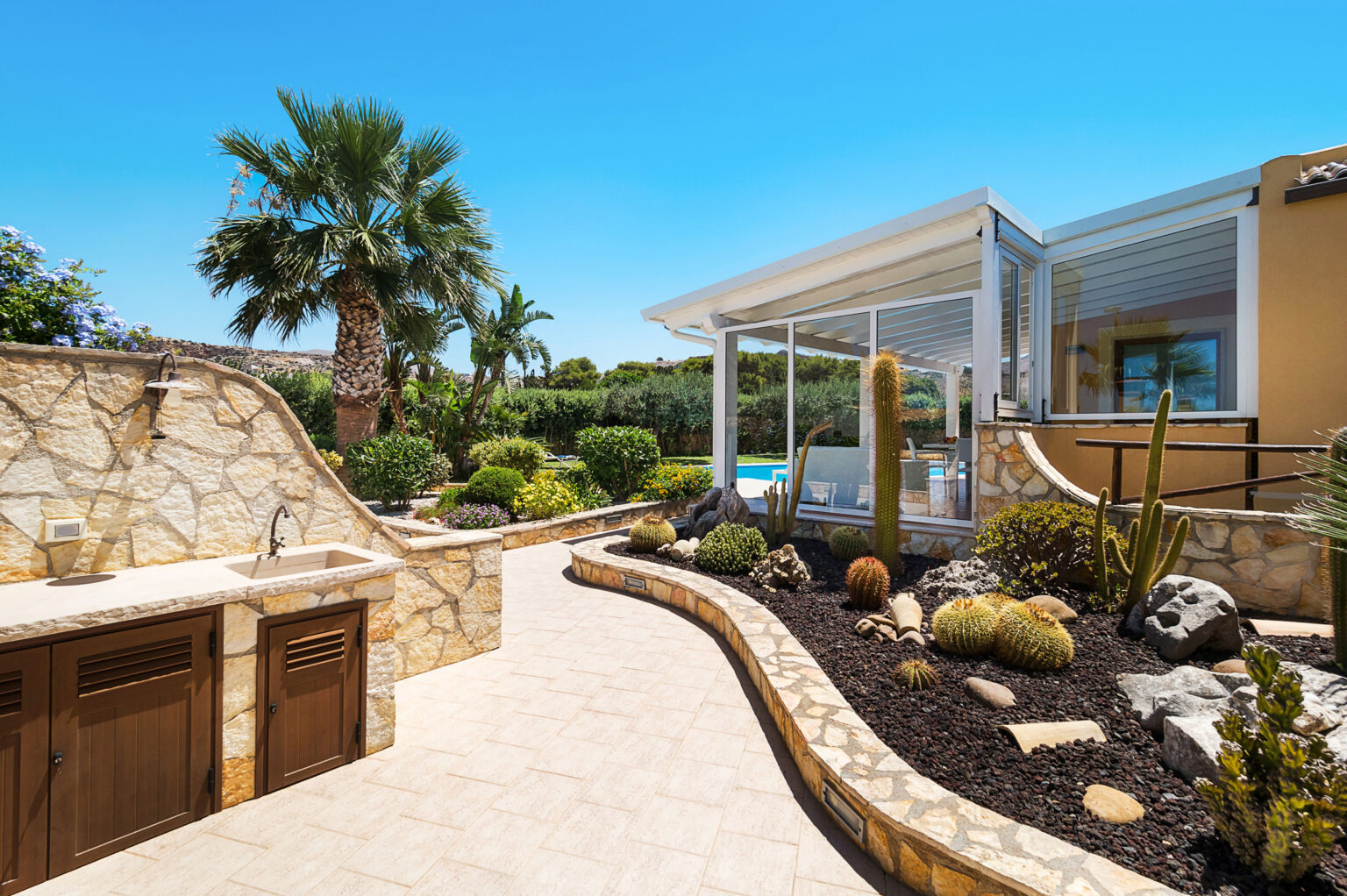 Stunning outdoor patio featuring a stone pathway, lush greenery, and cacti, with a modern glass-enclosed area overlooking a swimming pool under a clear blue sky. | Only on SopranoVillas