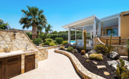 Stunning outdoor patio featuring a stone pathway, lush greenery, and cacti, with a modern glass-enclosed area overlooking a swimming pool under a clear blue sky.