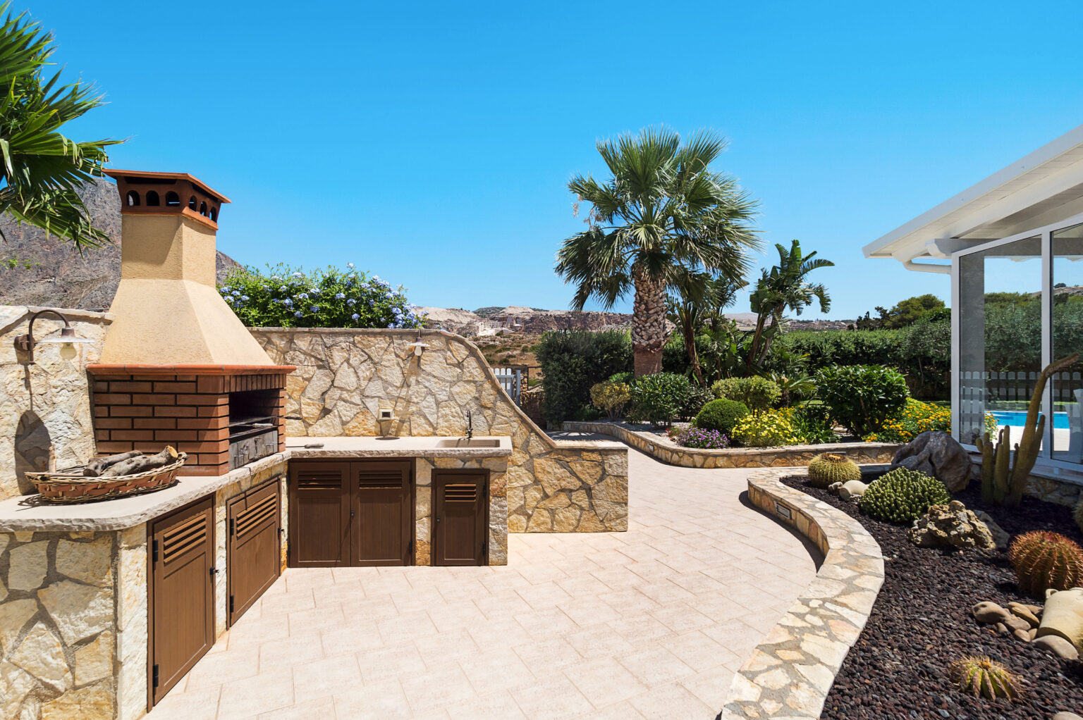 Outdoor kitchen with stone barbecue and cabinets, surrounded by lush greenery and palm trees, under a clear blue sky. Ideal for entertaining and outdoor dining. | Only on SopranoVillas