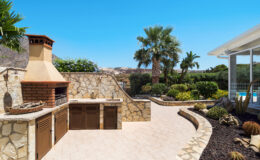 Outdoor kitchen with stone barbecue and cabinets, surrounded by lush greenery and palm trees, under a clear blue sky. Ideal for entertaining and outdoor dining.