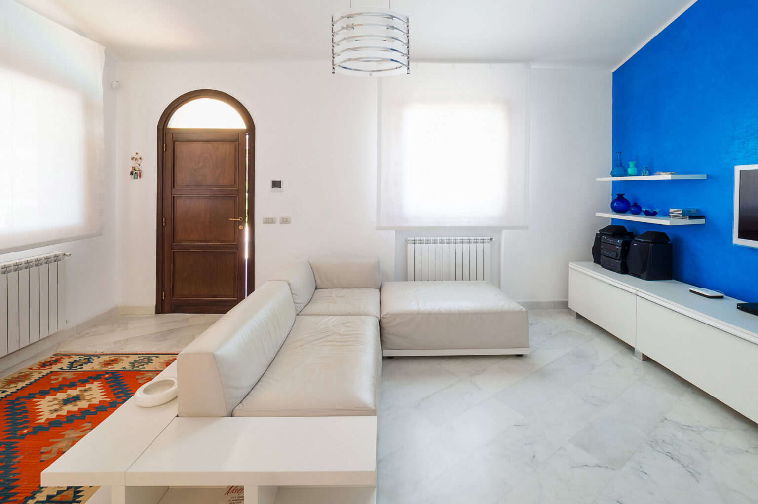 Modern living room featuring a light-colored sectional sofa, a vibrant blue accent wall, and a minimalist white entertainment unit. Natural light streams through sheer curtains, illuminating the marble flooring and decorative elements like a colorful area rug and decorative glass items on shelves. | Only on SopranoVillas