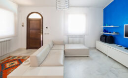 Modern living room featuring a light-colored sectional sofa, a vibrant blue accent wall, and a minimalist white entertainment unit. Natural light streams through sheer curtains, illuminating the marble flooring and decorative elements like a colorful area rug and decorative glass items on shelves.