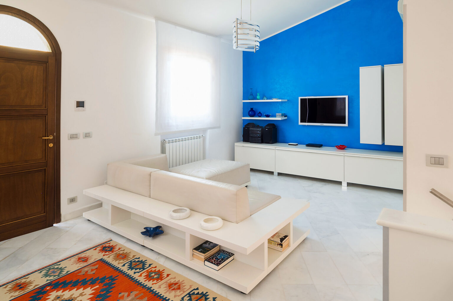 Modern living room with white furniture, a blue accent wall, and a flat-screen TV. Features include a stylish coffee table and decorative rug, creating a bright and inviting atmosphere. | Only on SopranoVillas