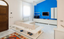 Modern living room with white furniture, a blue accent wall, and a flat-screen TV. Features include a stylish coffee table and decorative rug, creating a bright and inviting atmosphere.