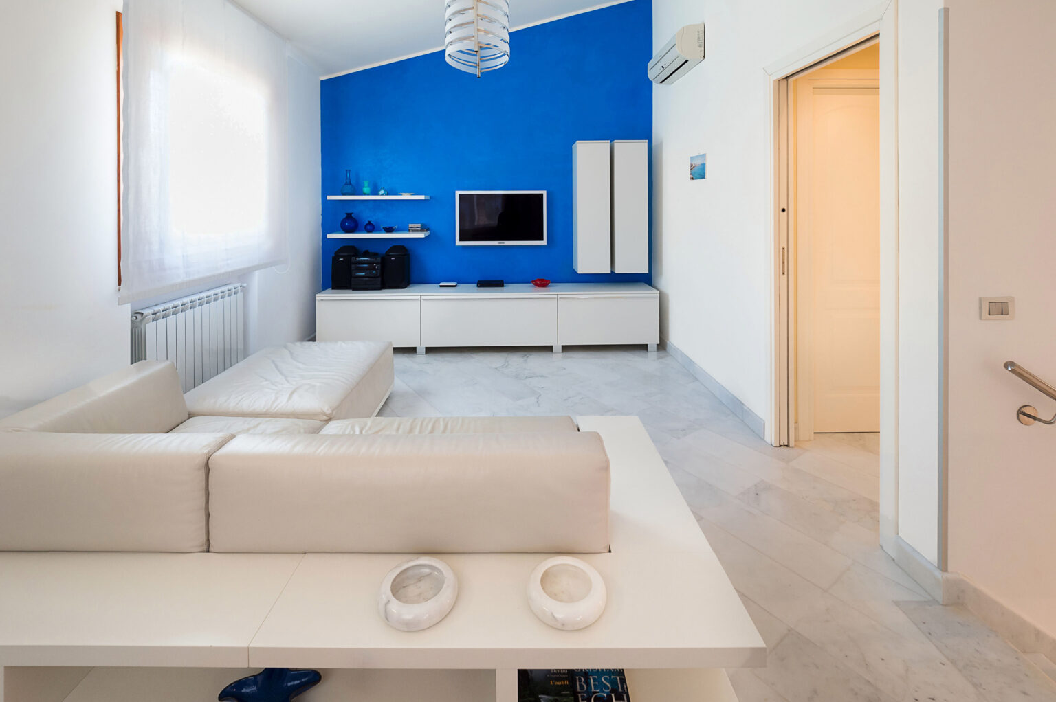 Modern living room featuring a white leather sofa, a vibrant blue accent wall, and minimalist furniture. The space includes a flat-screen TV mounted on a white media console, decorative glass vases on shelves, and natural light streaming through sheer curtains, creating an open and airy atmosphere. | Only on SopranoVillas