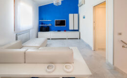 Modern living room featuring a white leather sofa, a vibrant blue accent wall, and minimalist furniture. The space includes a flat-screen TV mounted on a white media console, decorative glass vases on shelves, and natural light streaming through sheer curtains, creating an open and airy atmosphere.