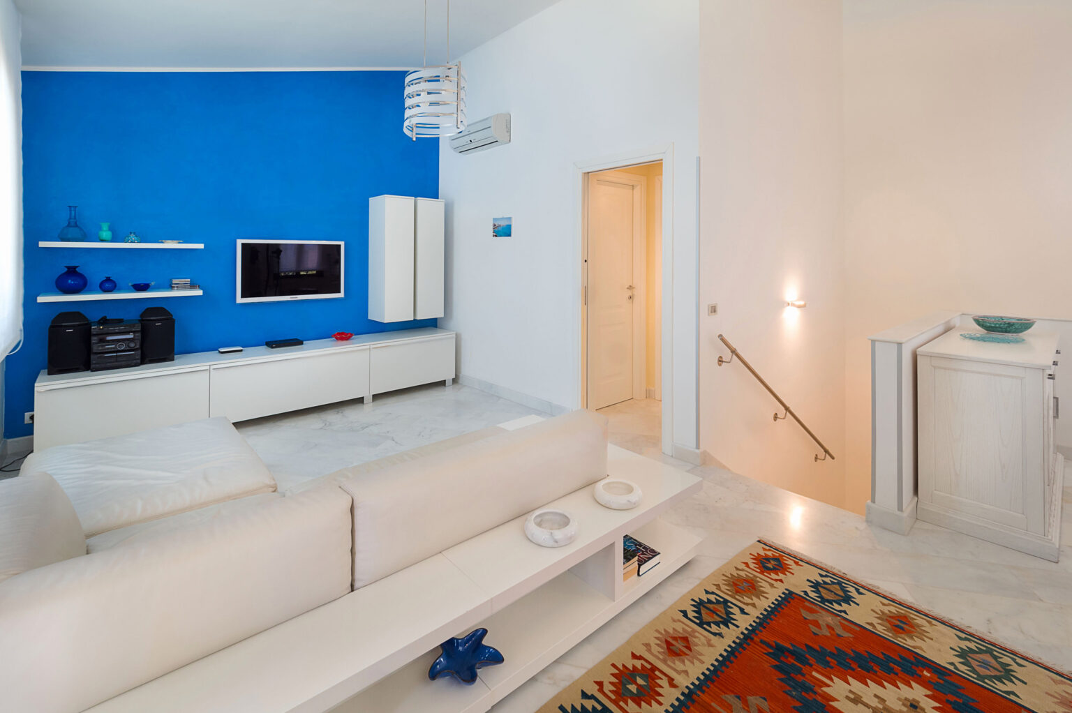Modern living room interior featuring a striking blue accent wall, white furniture, and a cozy rug. The space includes a television unit, decorative shelves, and soft lighting, creating a stylish and inviting atmosphere. | Only on SopranoVillas