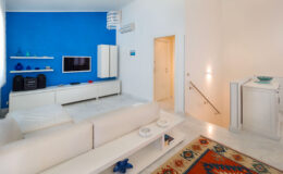 Modern living room interior featuring a striking blue accent wall, white furniture, and a cozy rug. The space includes a television unit, decorative shelves, and soft lighting, creating a stylish and inviting atmosphere.