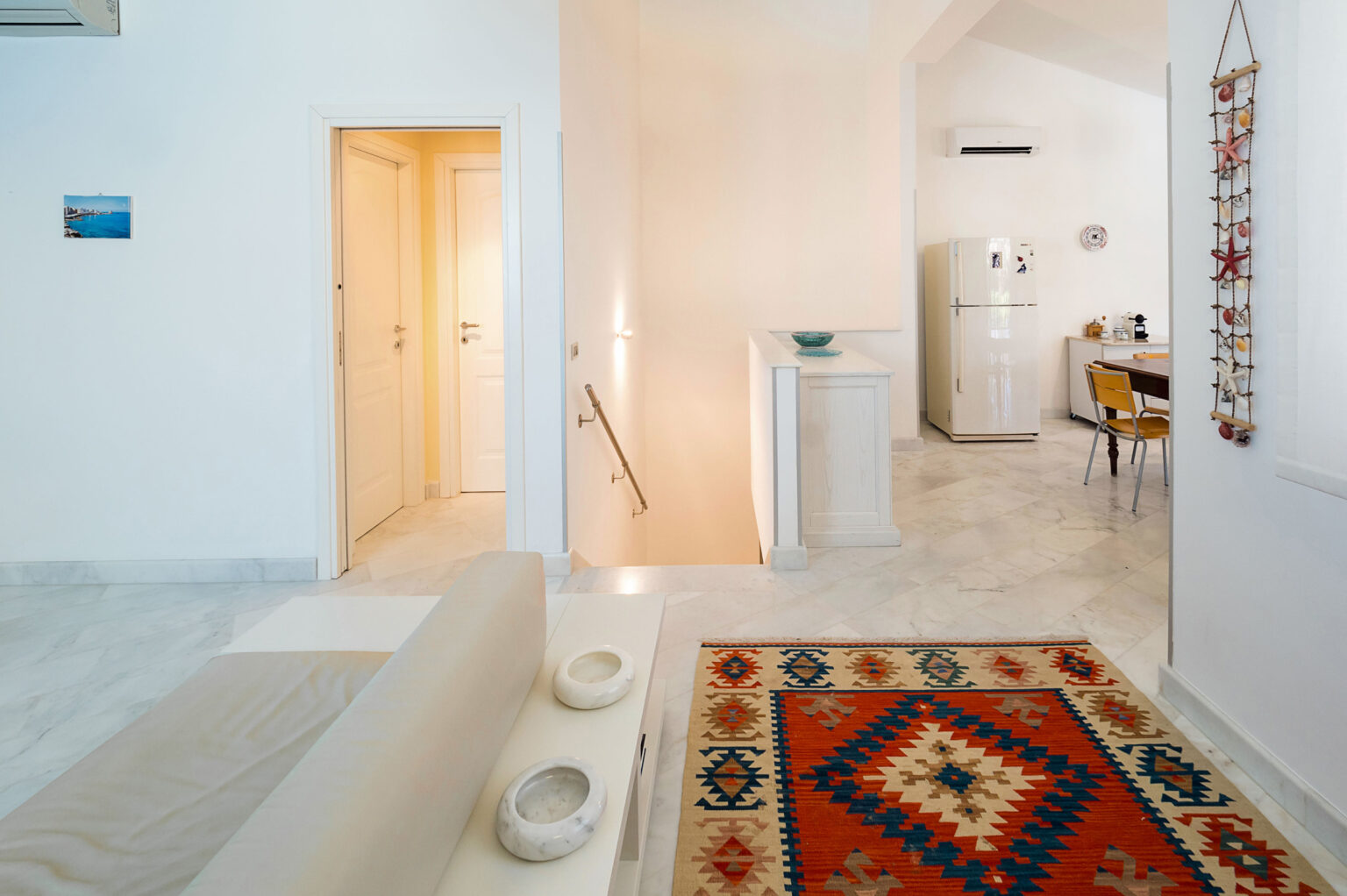 Bright and modern interior of a stylish living space featuring a cozy seating area, colorful patterned rug, and a dining area with a table and chairs. The open layout includes a view of the kitchen and a staircase leading to upper levels, complemented by white walls and marble flooring. | Only on SopranoVillas