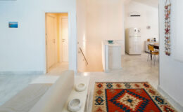 Bright and modern interior of a stylish living space featuring a cozy seating area, colorful patterned rug, and a dining area with a table and chairs. The open layout includes a view of the kitchen and a staircase leading to upper levels, complemented by white walls and marble flooring.