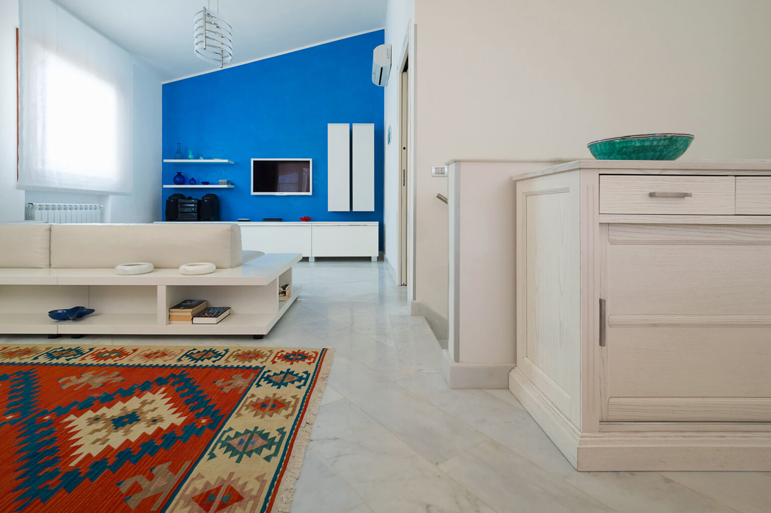 Modern living room interior featuring a blue accent wall, white furniture, and a decorative area rug. The space is bright and airy, with natural light streaming in through large windows. A sleek entertainment unit and minimalist décor complete the stylish design. | Only on SopranoVillas