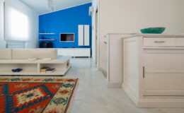 Modern living room interior featuring a blue accent wall, white furniture, and a decorative area rug. The space is bright and airy, with natural light streaming in through large windows. A sleek entertainment unit and minimalist décor complete the stylish design.