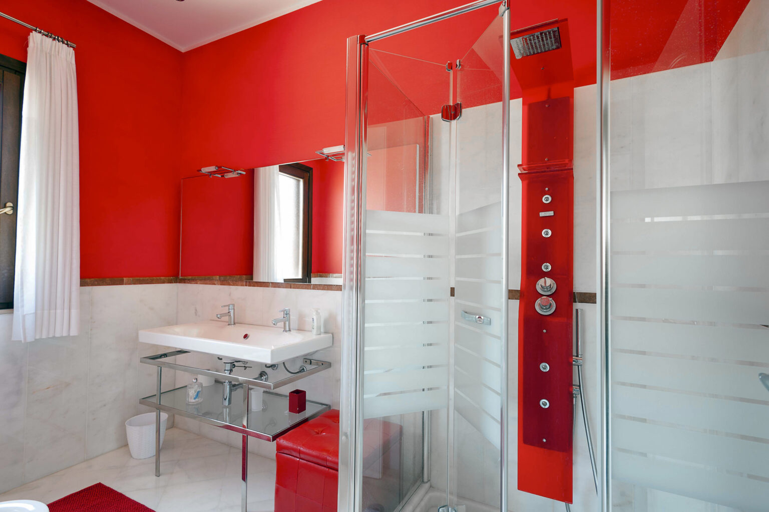 Modern bathroom featuring a vibrant red accent wall, a glass shower enclosure, a sleek glass sink vanity, and contemporary fixtures, creating a stylish and inviting space. | Only on SopranoVillas