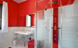 Modern bathroom featuring a vibrant red accent wall, a glass shower enclosure, a sleek glass sink vanity, and contemporary fixtures, creating a stylish and inviting space.