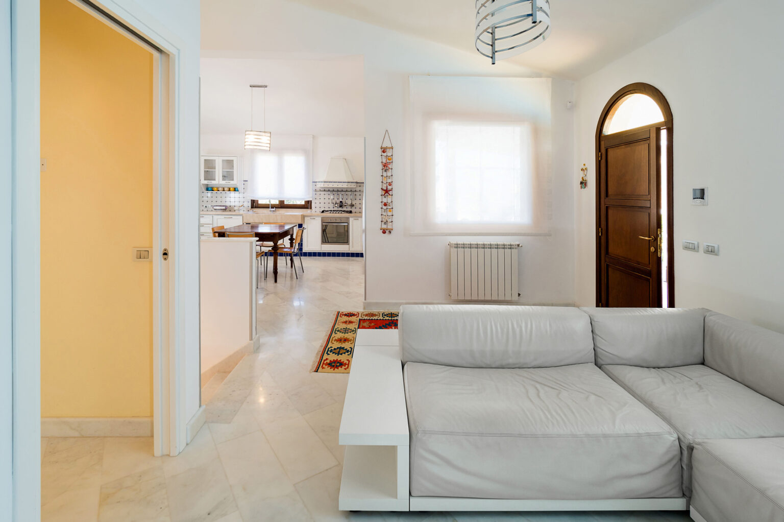 Bright and spacious living room featuring a light gray sofa, an arched wooden door, and an open view into a modern kitchen. The room has white walls and ample natural light from large windows, creating a welcoming atmosphere. | Only on SopranoVillas
