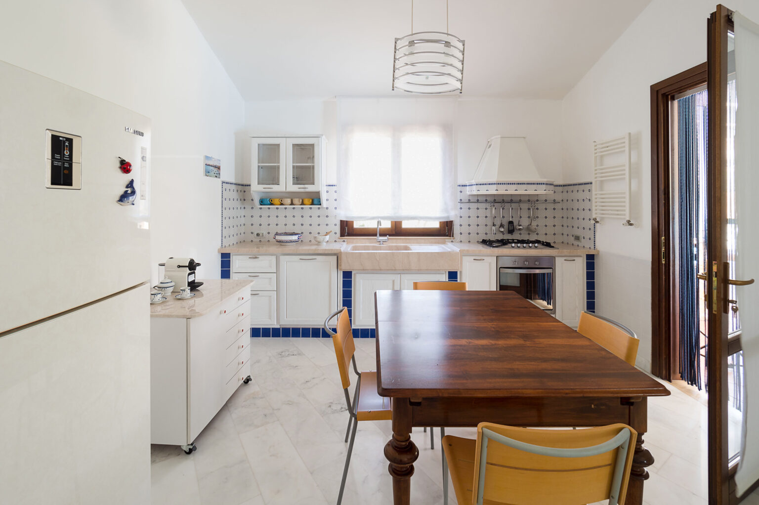 Bright and airy kitchen featuring a wooden dining table surrounded by yellow chairs, modern appliances, and white cabinetry with blue tile accents. Natural light floods the space through a large window. | Only on SopranoVillas