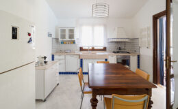 Bright and airy kitchen featuring a wooden dining table surrounded by yellow chairs, modern appliances, and white cabinetry with blue tile accents. Natural light floods the space through a large window.