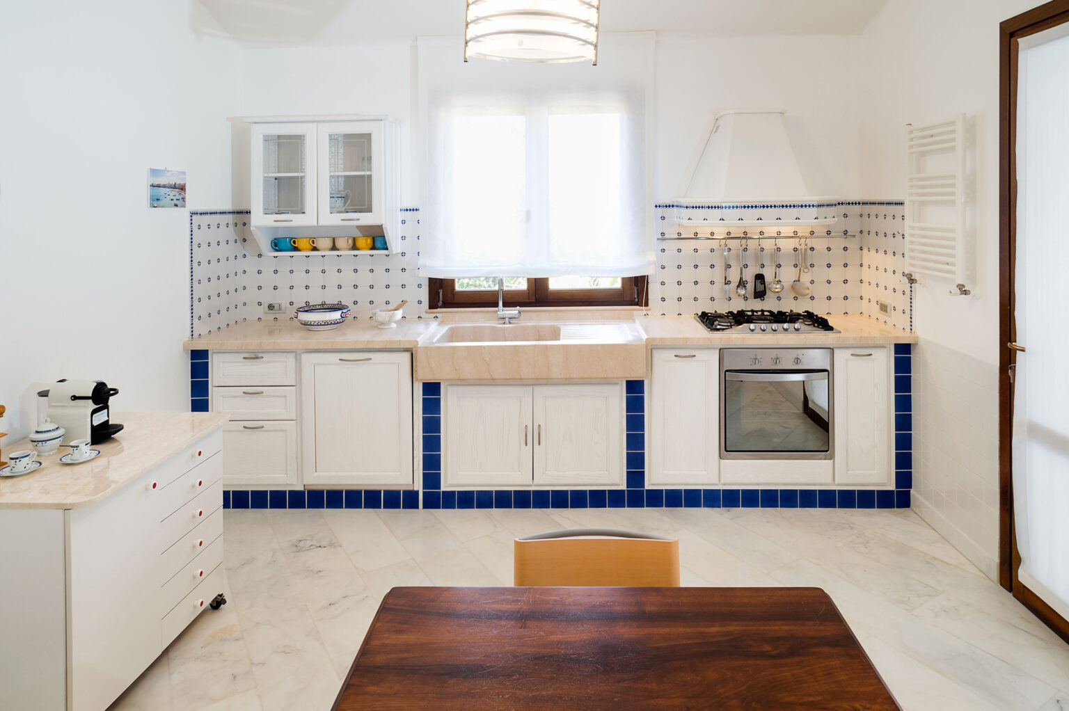 Modern kitchen interior featuring white cabinetry, blue tiled accents, and a wooden dining table. The space includes a sink, stove, and ample natural light from a window. Ideal for home cooking and entertaining. | Only on SopranoVillas