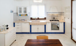 Modern kitchen interior featuring white cabinetry, blue tiled accents, and a wooden dining table. The space includes a sink, stove, and ample natural light from a window. Ideal for home cooking and entertaining.