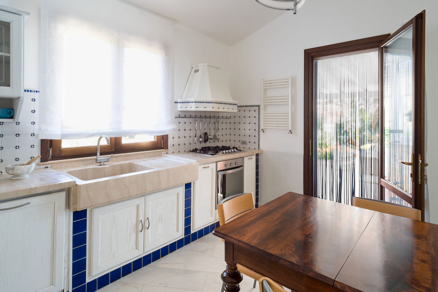 Bright and airy kitchen featuring a large sink, wooden dining table, and tiled backsplash, with natural light streaming through the windows and a view of the outdoor patio. | Only on SopranoVillas
