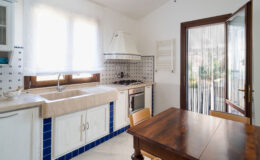 Bright and airy kitchen featuring a large sink, wooden dining table, and tiled backsplash, with natural light streaming through the windows and a view of the outdoor patio.