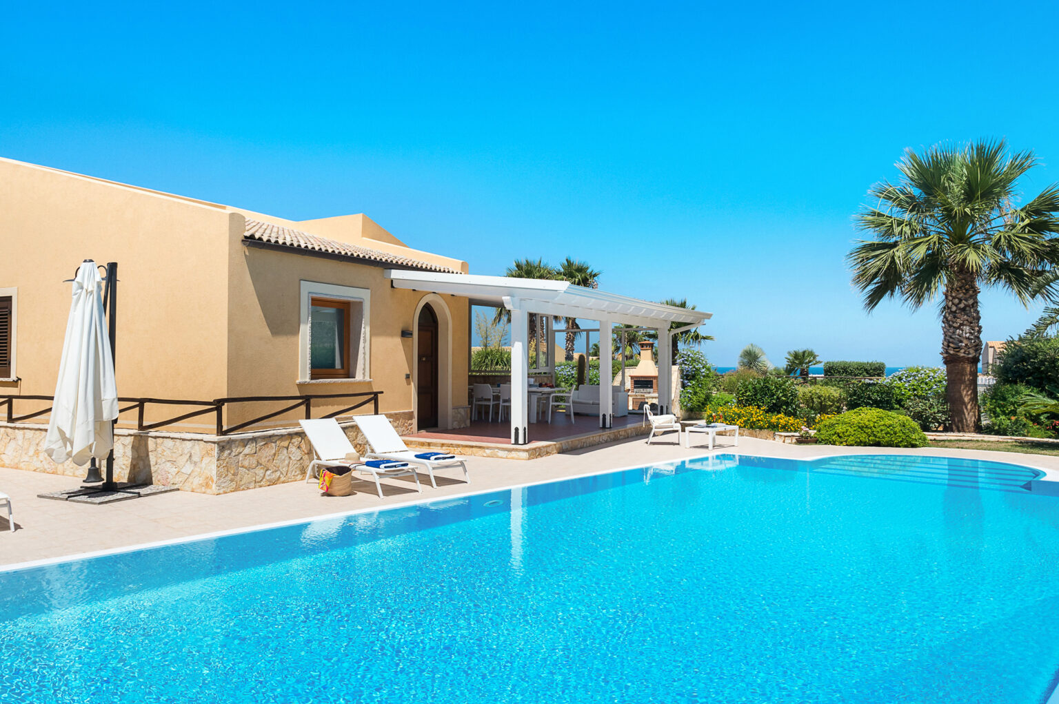 Modern villa with a private swimming pool, surrounded by palm trees and lush greenery under a clear blue sky. Ideal for luxury vacation rentals. | Only on SopranoVillas