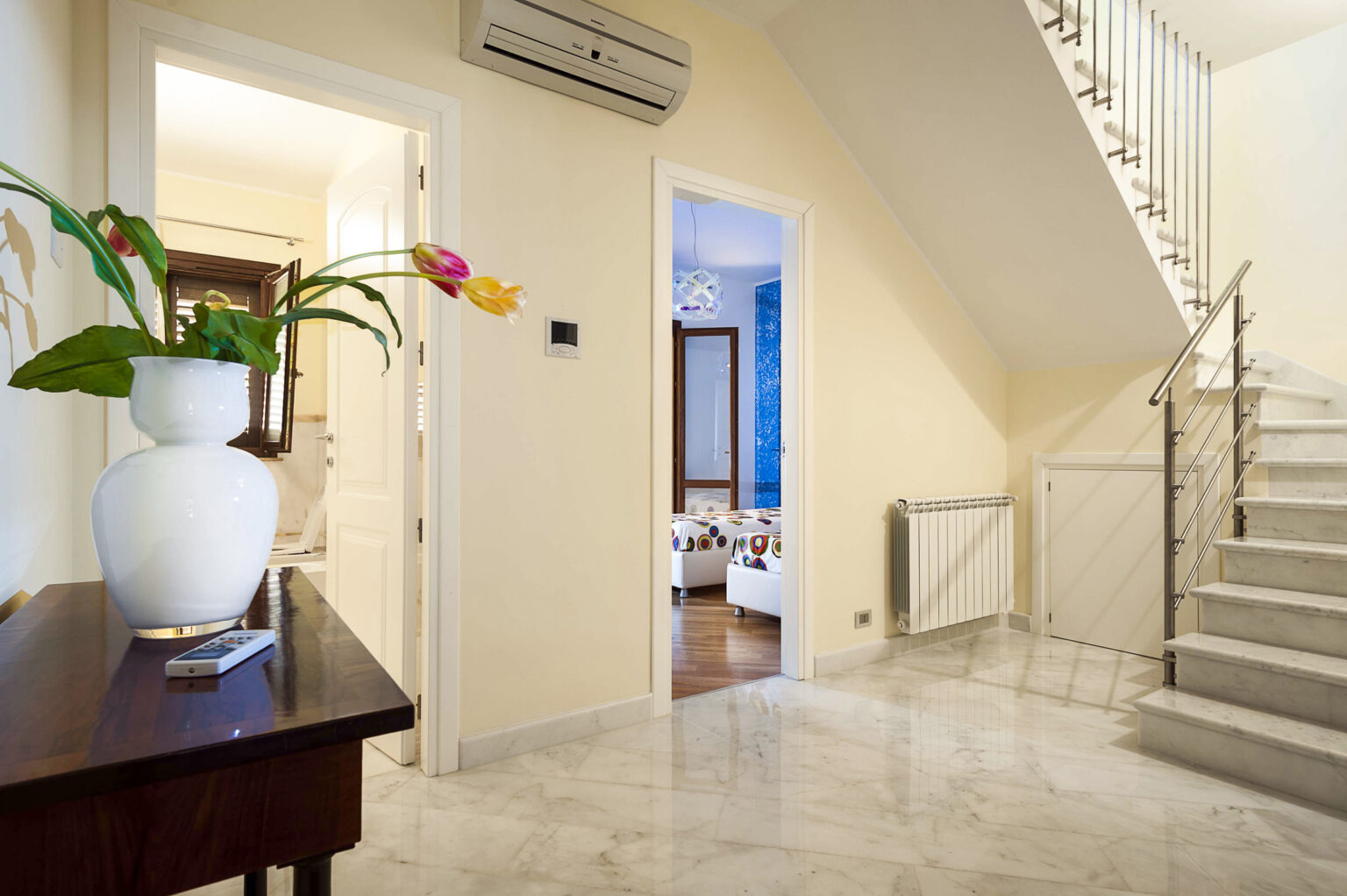 Modern interior hallway featuring a stylish staircase, marble flooring, and a decorative vase with flowers. The space is well-lit, showcasing a warm color palette and contemporary decor elements. | Only on SopranoVillas