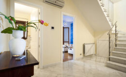 Modern interior hallway featuring a stylish staircase, marble flooring, and a decorative vase with flowers. The space is well-lit, showcasing a warm color palette and contemporary decor elements.