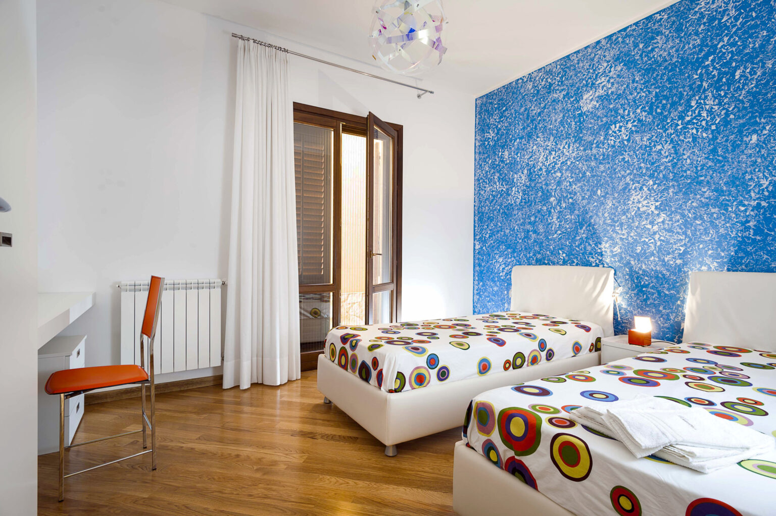Cozy bedroom featuring two twin beds with colorful patterned bedspreads, a modern chair, and a vibrant blue accent wall. Natural light streams through the open door, enhancing the warm wooden floor. Ideal for relaxation and comfort. | Only on SopranoVillas