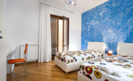 Cozy bedroom featuring two twin beds with colorful patterned bedspreads, a modern chair, and a vibrant blue accent wall. Natural light streams through the open door, enhancing the warm wooden floor. Ideal for relaxation and comfort.