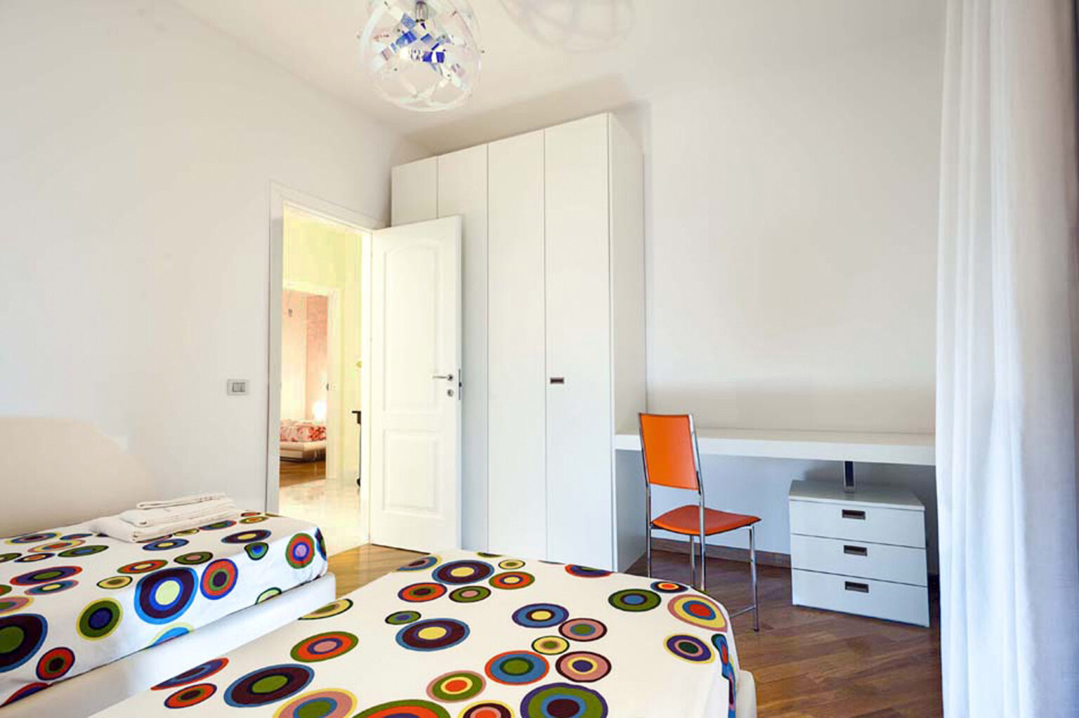 Bright and modern bedroom featuring two beds with colorful patterned bedding, a white wardrobe, a desk with a chair, and hardwood flooring. The room is well-lit and has a door leading to another space. | Only on SopranoVillas