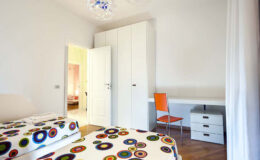 Bright and modern bedroom featuring two beds with colorful patterned bedding, a white wardrobe, a desk with a chair, and hardwood flooring. The room is well-lit and has a door leading to another space.
