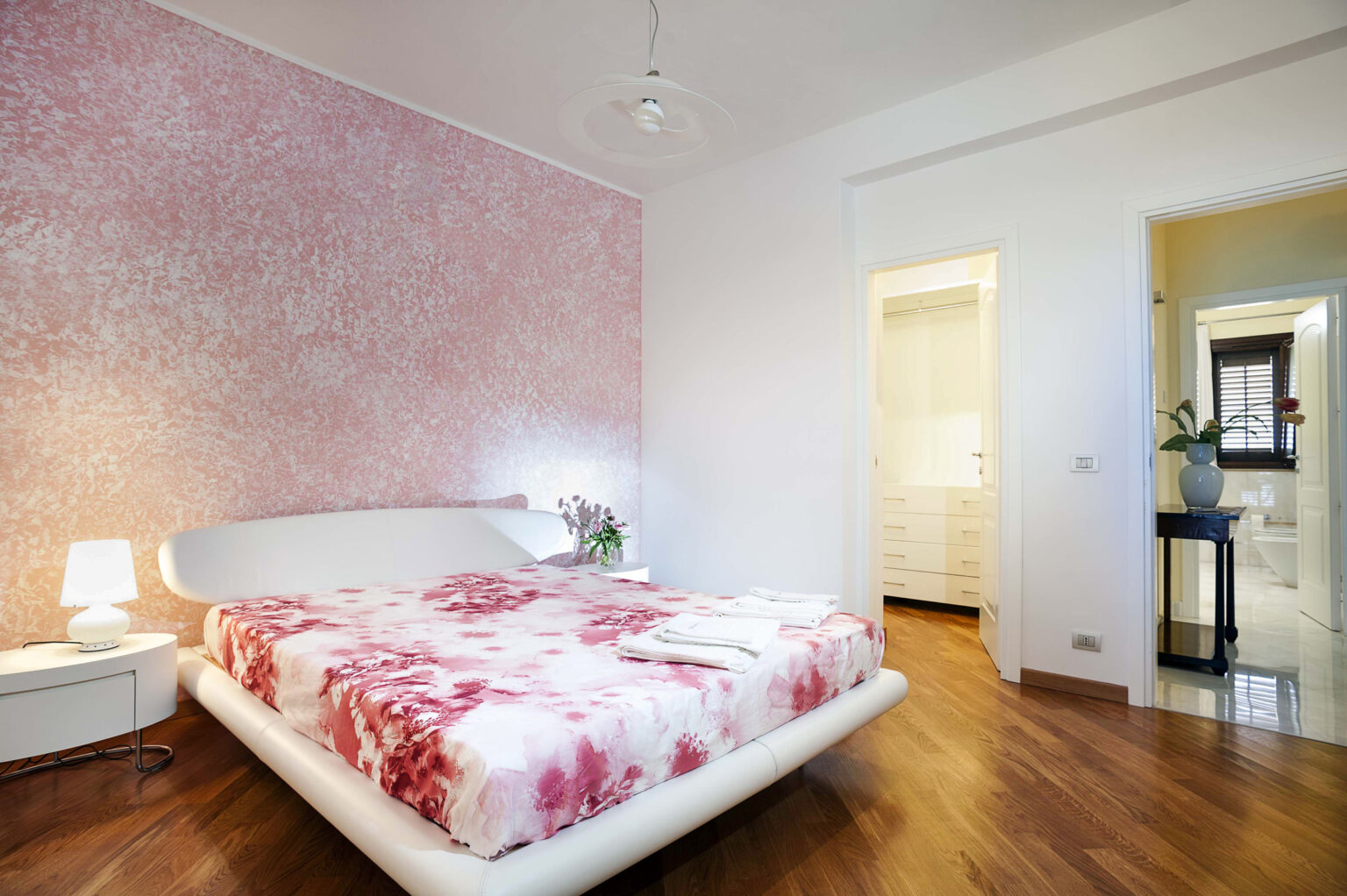 Modern bedroom featuring a stylish white bed with a pink floral bedspread, soft lighting from a bedside lamp, and a textured pink wall. The room includes a doorway leading to an en-suite bathroom and a closet area, with polished wooden flooring enhancing the contemporary design. | Only on SopranoVillas